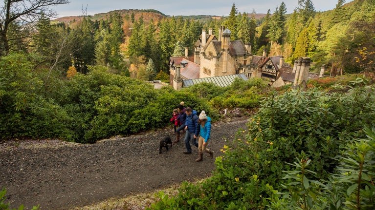 A family, 3 adults, 1 child and a black dog, are walking along a path at Cragside. The chimneys of Cragside House are visible. The day is cold and wintry. Everyone is wearing bobble hats.
